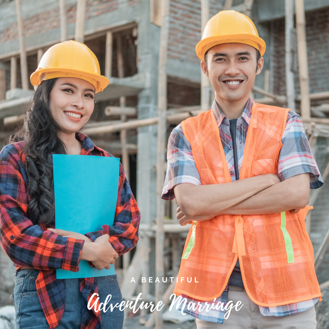 A couple at a construction site, both are wearing hard hats. The lady is holding blueprints. They are both smiling.